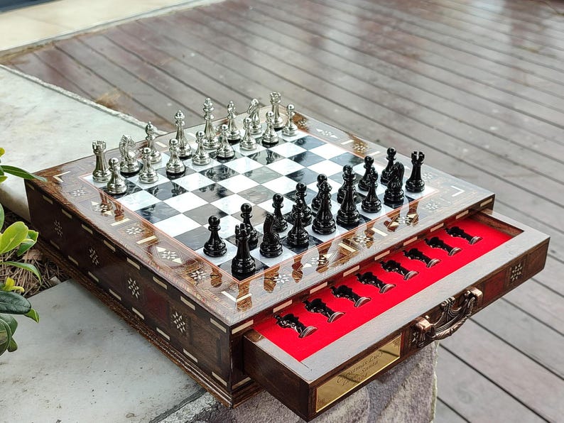 K&ouml;nnte beinhalten: Ein Schachspiel aus Holz mit einer Klappbox. Das Schachbrett ist mit Perlmutt eingelegt und die Figuren sind silber und schwarz. Die Box hat ein rotes Filz-Innenfutter und einen Messinggriff.