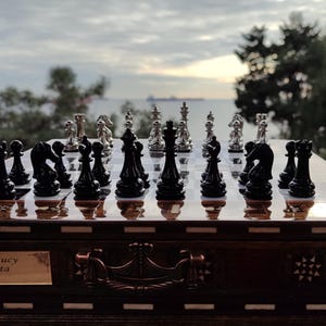 K&ouml;nnte beinhalten: Ein Schachbrett aus Holz mit einem silbernen und schwarzen Schachspiel. Das Schachbrett hat eine dunkelbraune Holzoptik und ist mit aufwendigen Schnitzereien verziert. Die Schachfiguren sind in ihren Startpositionen aufgestellt. Das Schachbrett steht auf einem Tisch vor einem Fenster mit Blick auf das Meer. Der Text "Christmas Lucy Love, Santa 2024" ist in das Schachbrett eingraviert.