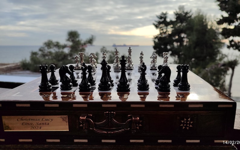K&ouml;nnte beinhalten: Ein Schachbrett aus Holz mit einem silbernen und schwarzen Schachspiel. Das Schachbrett hat eine dunkelbraune Holzoptik und ist mit aufwendigen Schnitzereien verziert. Die Schachfiguren sind in ihren Startpositionen aufgestellt. Das Schachbrett steht auf einem Tisch vor einem Fenster mit Blick auf das Meer. Der Text "Christmas Lucy Love, Santa 2024" ist in das Schachbrett eingraviert.