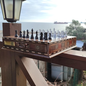 K&ouml;nnte beinhalten: Ein Schachbrett aus Holz mit einer Schublade und eingelegten Details. Das Brett ist mit schwarzen und wei&szlig;en Schachfiguren aufgestellt. Das Brett steht auf einem Holzgel&auml;nder mit Blick auf das Meer im Hintergrund.