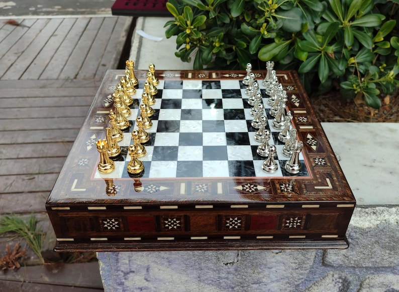 K&ouml;nnte beinhalten: Ein Schachbrett aus Holz mit eingelegten Details und einer schwarz-wei&szlig;en Schachbrettfl&auml;che. Die Schachfiguren sind golden und silbern.