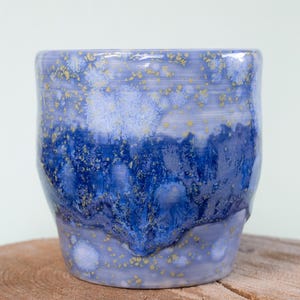 May include: A ceramic mug featuring a multi-toned blue glaze with a crystalline texture. The upper portion is a lighter periwinkle blue, transitioning into a deep, saturated cobalt blue in the center, with small gold-colored speckles throughout.
