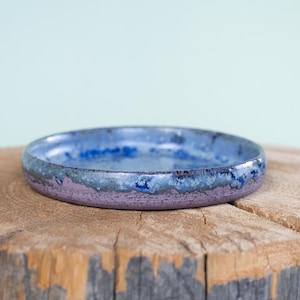 May include: A shallow, circular ceramic dish featuring a reactive glaze with a deep blue interior that transitions into a mottled blue and purple exterior. The rim shows a textured, crystalline finish where the blue and purple glazes overlap.