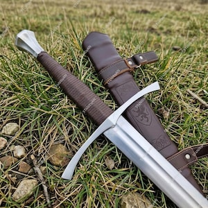 May include: A medieval sword with a silver blade and guard, a brown leather-wrapped handle, and a matching leather sheath. The sword rests on a bed of green grass, showcasing its craftsmanship and historical design.
