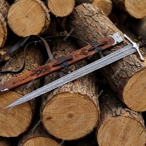 Puede incluir: Espada y vaina sobre troncos de madera. La espada tiene una hoja plateada con diseño, empuñadura marrón y guarda plateada. La vaina de madera y cuero, con grabados decorativos, muestra artesanía.