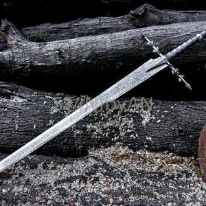 May include: A silver sword with a textured blade and a black handle, lying on dark wooden logs. The sword has a crossguard with decorative elements and a pointed tip, evoking a medieval or fantasy theme.