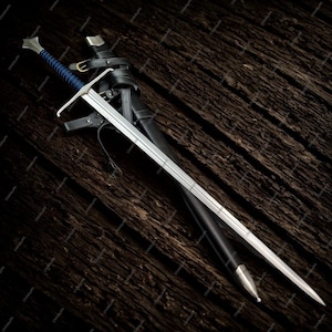 May include: A long, silver sword with a blue handle and a black leather sheath. The sword has a crossguard and a pointed tip. The sheath has a silver tip and is secured with black leather straps. The sword rests on a dark wooden surface.