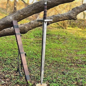 May include: A long, silver sword with a brown leather-wrapped handle and a crossguard. The sword is displayed with its brown leather sheath, resting against a tree branch. The background is a grassy area with trees.