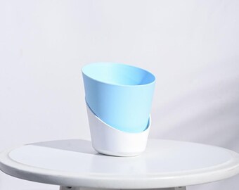 Puede incluir: Una maceta moderna de dos tonos, con una sección superior azul claro y una base blanca. La maceta tiene un diseño angular único y se encuentra sobre una mesa blanca. El fondo es blanco liso.
