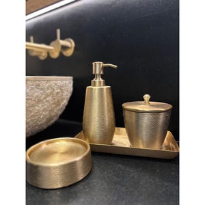 May include: A gold-toned bathroom set comprising a soap dispenser, a small lidded jar, and a soap dish, each adorned with a wave pattern. The items are arranged on a rectangular tray, offering a coordinated and stylish appearance.