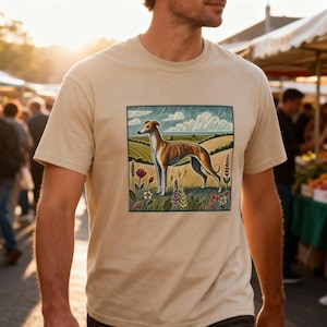 May include: Beige t-shirt with a graphic of a greyhound dog in a field. The dog is brown and white, set against a backdrop of green fields and a cloudy sky. The shirt is made of a soft, neutral-coloured fabric. The image is framed with a black border.