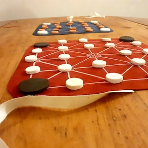 Puede incluir: Dos juegos de mesa se muestran sobre una superficie de madera. El tablero principal es rojo con marcas blancas y piezas blancas y negras. Un segundo tablero, azul con piezas naranjas y blancas, es visible en el fondo. Una cinta blanca está en primer plano.