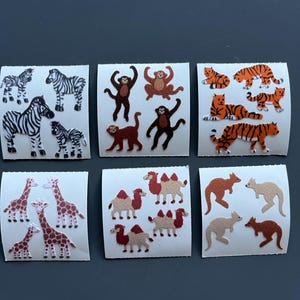 Könnte beinhalten: Eine Sammlung von Tieraufklebern mit Zebras, Affen, Tigern, Giraffen, Kamelen und Kängurus. Die Aufkleber sind in verschiedenen Farben wie Schwarz und Weiß, Braun, Orange und Beige gehalten und auf weißen rechteckigen Hintergründen angeordnet.