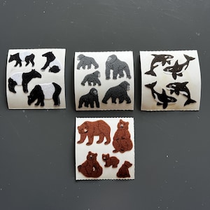 Pode incluir: Um conjunto de autocolantes com tema animal, apresentando pandas, gorilas, ursos e orcas. Os autocolantes são em preto, branco, castanho e cinzento, com uma textura. Os autocolantes estão sobre um fundo branco.