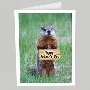 May include: A groundhog standing on its hind legs in a grassy field holding a wooden sign that says "Happy Father's Day".