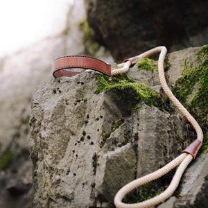 May include: A brown leather camera strap with a beige rope attached to it. The strap is lying on a rocky surface covered in moss.