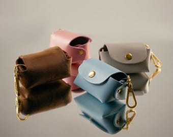 May include: Four small leather pouches in brown, pink, blue, and gray, each with a gold-colored clasp. The pouches are rectangular with a curved flap and a gold snap closure. They are arranged on a reflective surface.