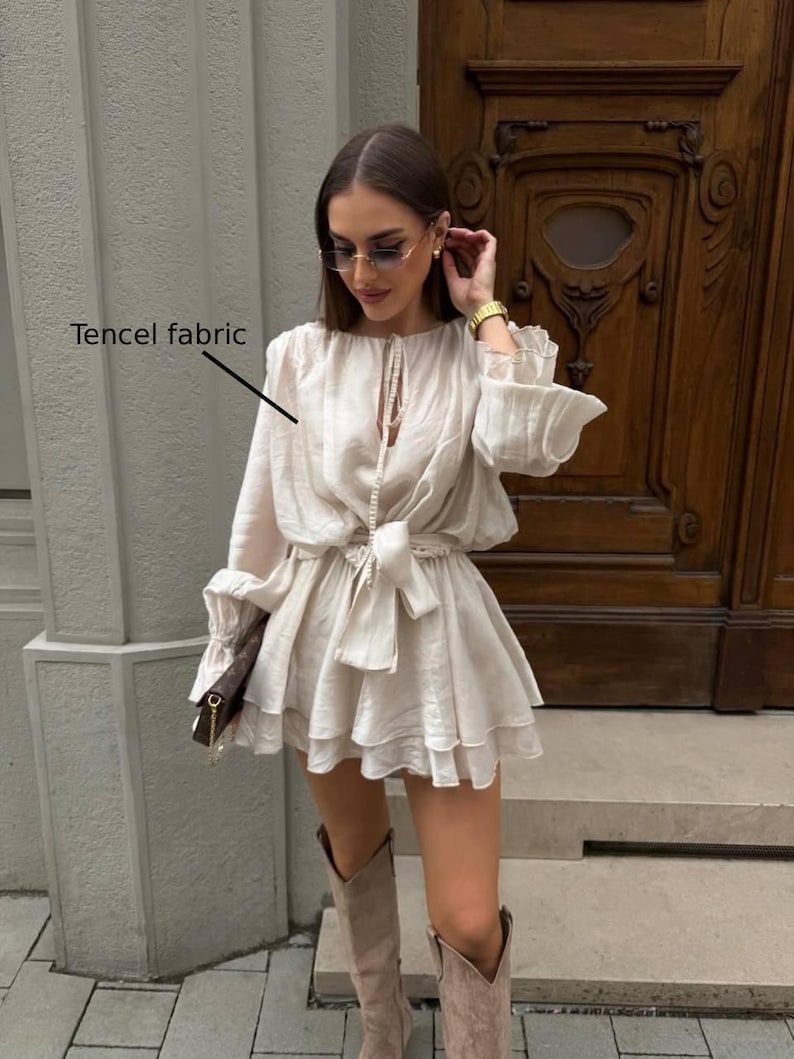 May include: A cream-colored Tencel fabric dress with long sleeves and a tiered skirt. The dress features a tie-waist and is paired with knee-high boots. The image shows the dress in a natural light setting.