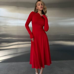 May include: A long-sleeved, red dress with a flared skirt. The dress has a round neckline and fitted bodice. The model is wearing white strappy heels. The background is a metallic silver wall.