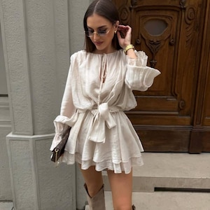 May include: A cream-colored romper with long sleeves and a tiered skirt, cinched at the waist with a matching tie. The outfit is paired with knee-high boots and sunglasses. A small brown purse is held in the hand.