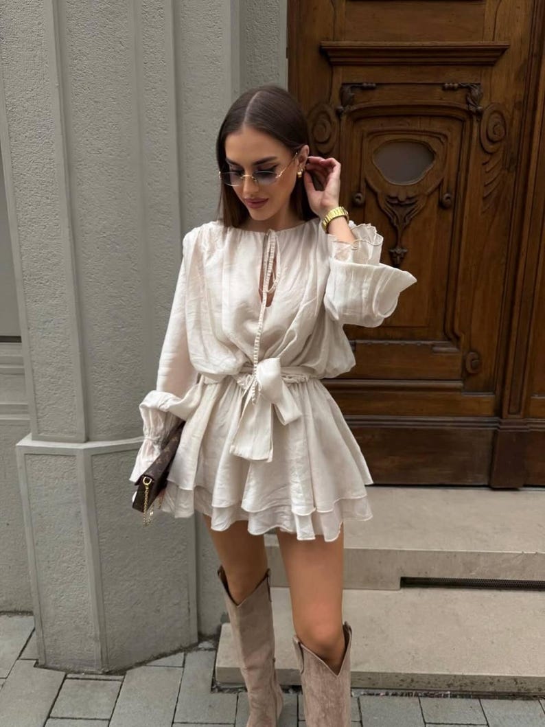 May include: A cream-colored romper with long sleeves and a tiered skirt, cinched at the waist with a matching tie. The outfit is paired with knee-high boots and sunglasses. A small brown purse is held in the hand.