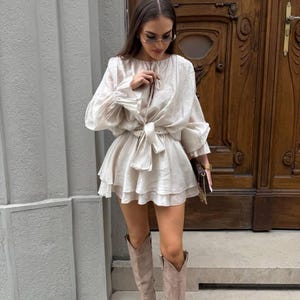 May include: A beige dress with long sleeves and a tiered skirt. The dress is cinched at the waist with a matching belt. The outfit is paired with knee-high, light brown suede boots. The background features a wooden door and a gray wall.