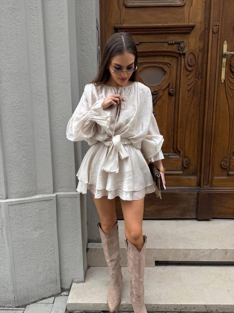 May include: A beige dress with long sleeves and a tiered skirt. The dress is cinched at the waist with a matching belt. The outfit is paired with knee-high, light brown suede boots. The background features a wooden door and a gray wall.