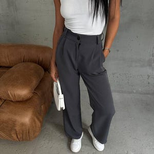 Puede incluir: Una mujer con un top blanco de tirantes y pantalones grises de pierna ancha con un botón blanco. Lleva un bolso blanco y zapatillas blancas.