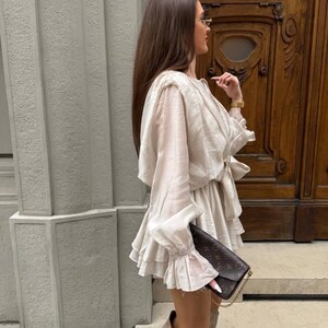 May include: A cream-colored dress with ruffled layers and long sleeves with ruffled cuffs. The dress is paired with brown suede boots and a brown patterned clutch. The background features a textured wall and a wooden door.
