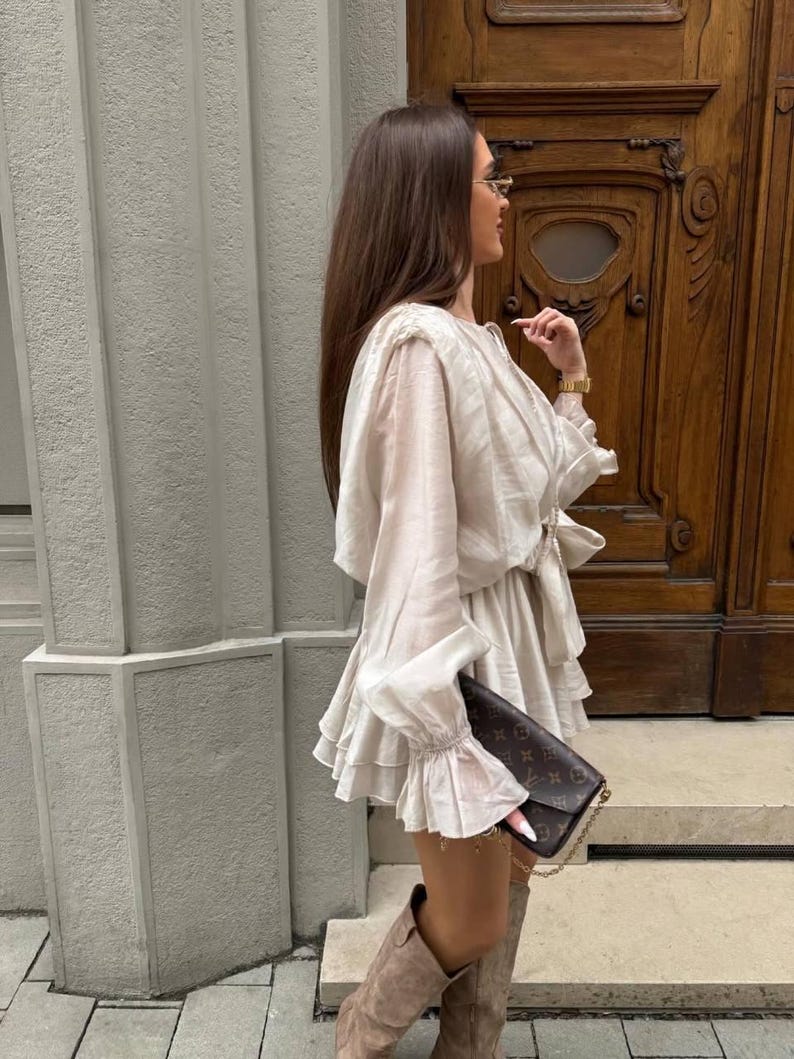 May include: A cream-colored dress with ruffled layers and long sleeves with ruffled cuffs. The dress is paired with brown suede boots and a brown patterned clutch. The background features a textured wall and a wooden door.