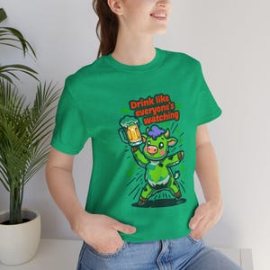 May include: Green t-shirt featuring a cartoon green cow holding a beer mug. The text on the shirt reads "Drink like everyone's watching".