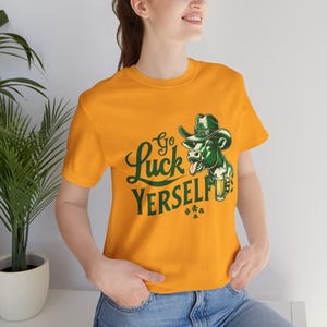 May include: Mustard yellow t-shirt with a green cartoon cow wearing a cowboy hat and holding a beer. The shirt has the text "Go Luck Yerself" in green script, with shamrock accents.