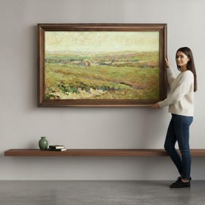 Puede incluir: Una pintura de paisaje enmarcada en un marco de madera marrón. La obra de arte representa un campo con tonos verdes y amarillos. Una mujer sostiene la pintura. Un jarrón verde y dos libros están en una estantería de madera debajo.