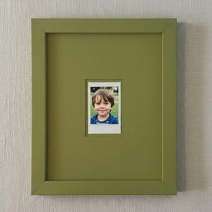 May include: A square, olive green picture frame with a white-bordered photograph of a smiling child. The frame has a simple, modern design and is mounted on a textured, off-white wall.