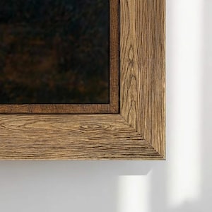 May include: A rectangular wooden framed mirror. The frame is made of light brown wood with visible grain. The mirror's surface is dark, reflecting a blurred image. The frame's corner is visible against a white background.