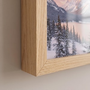 Puede incluir: Un marco de fotos de madera clara con una fotografía de paisaje. El marco tiene un diseño sencillo con un acabado de veta de madera natural. La fotografía muestra una escena de montaña nevada con árboles y un lago.