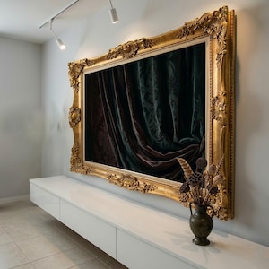 May include: A large, ornate gold picture frame with a dark, patterned curtain inside. The frame is highly detailed with intricate carvings. A small, dark vase with dried flowers sits below the frame on a white cabinet.