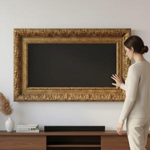 Può includere: Una grande cornice dorata ornata con interno nero è montata su una parete bianca. Una soundbar scura è posizionata su un mobile in legno sottostante. Una donna è in piedi vicino alla cornice.