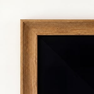 Puede incluir: Primer plano de un marco de madera con un acabado natural de vetas. El marco rodea una superficie negra, creando un contraste entre la madera clara y el interior oscuro. El marco está sobre un fondo blanco.