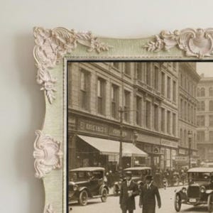 Pode incluir: Fotografia vintage a preto e branco de uma cena de rua da cidade, emoldurada numa moldura verde pálido com detalhes florais e de rolagem rosa claro. A fotografia apresenta carros clássicos e pessoas.