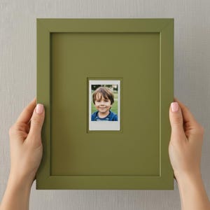 May include: A square, olive green picture frame holding a small photograph of a smiling child. The frame is held by two hands with pink nail polish. The photograph is centered within the frame.