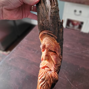 Puede incluir: Escultura de madera tallada a mano que representa la cara de un hombre barbudo. La escultura está hecha de madera marrón oscura, con la cara y la barba en un tono marrón anaranjado más claro. El tallado presenta detalles y texturas.