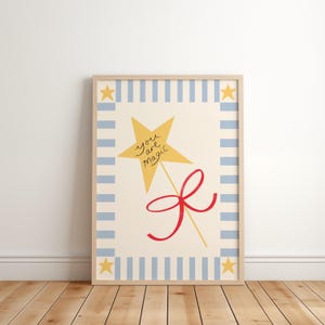 May include: A framed print with a yellow star wand and the words "you are magic". A red bow is tied below the wand. The print has a cream background with a blue striped border and yellow stars in each corner. The frame is light brown.