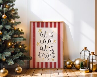 May include: A framed print with the text "all is calm all is bright" in a wooden frame. The print has a red and white striped border. A Christmas tree with gold ornaments and lights is to the left. Gold ornaments and candle holders are in front of the print.