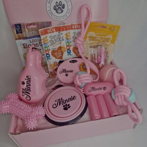 May include: Pink gift box filled with dog treats and toys, including a pink dog bowl, a pink rope toy, a pink chew toy, a pink retractable lead, and a bag of dog treats. The box is labelled "Minnie's Treats, Especially Fur You".