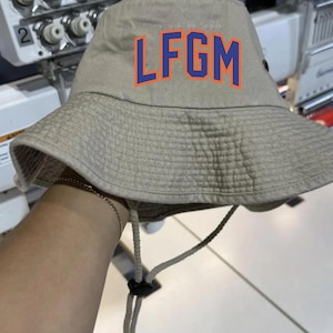 May include: A beige bucket hat featuring the letters "LFGM" in blue with an orange outline. The hat has a brim and an adjustable chin strap. The hat is made of a durable fabric, suitable for outdoor wear.