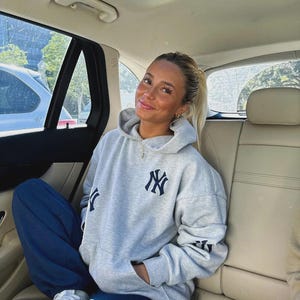 Puede incluir: Una sudadera gris claro con el logo de los Yankees de Nueva York en la parte delantera, la manga y la espalda. La persona lleva un pantal&oacute;n de ch&aacute;ndal azul marino y zapatillas blancas. La persona est&aacute; sentada en un coche.