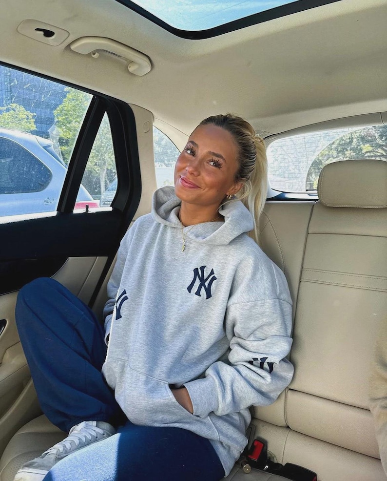 Puede incluir: Una sudadera gris claro con el logo de los Yankees de Nueva York en la parte delantera, la manga y la espalda. La persona lleva un pantal&oacute;n de ch&aacute;ndal azul marino y zapatillas blancas. La persona est&aacute; sentada en un coche.