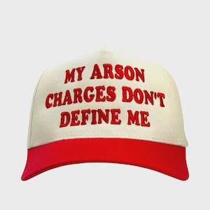 May include: A cream-colored baseball cap with a red brim. The cap features the embroidered text "MY ARSON CHARGES DON'T DEFINE ME" in red block letters. The hat is a classic six-panel design.