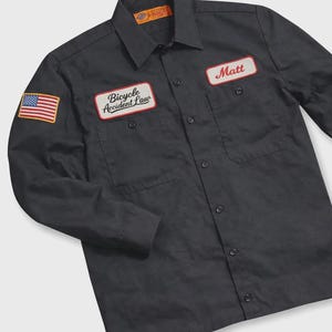 May include: Black work shirt with an American flag patch and two name patches. One patch reads "Matt" and the other says "Bicycle Accident Law". The shirt has two chest pockets and a button-down front.